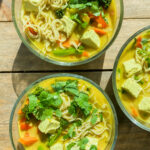 A steaming bowl of tofu curry noodle soup with colorful vegetables like red bell peppers and broccoli, garnished with fresh cilantro, served with udon noodles in a rich coconut curry broth.