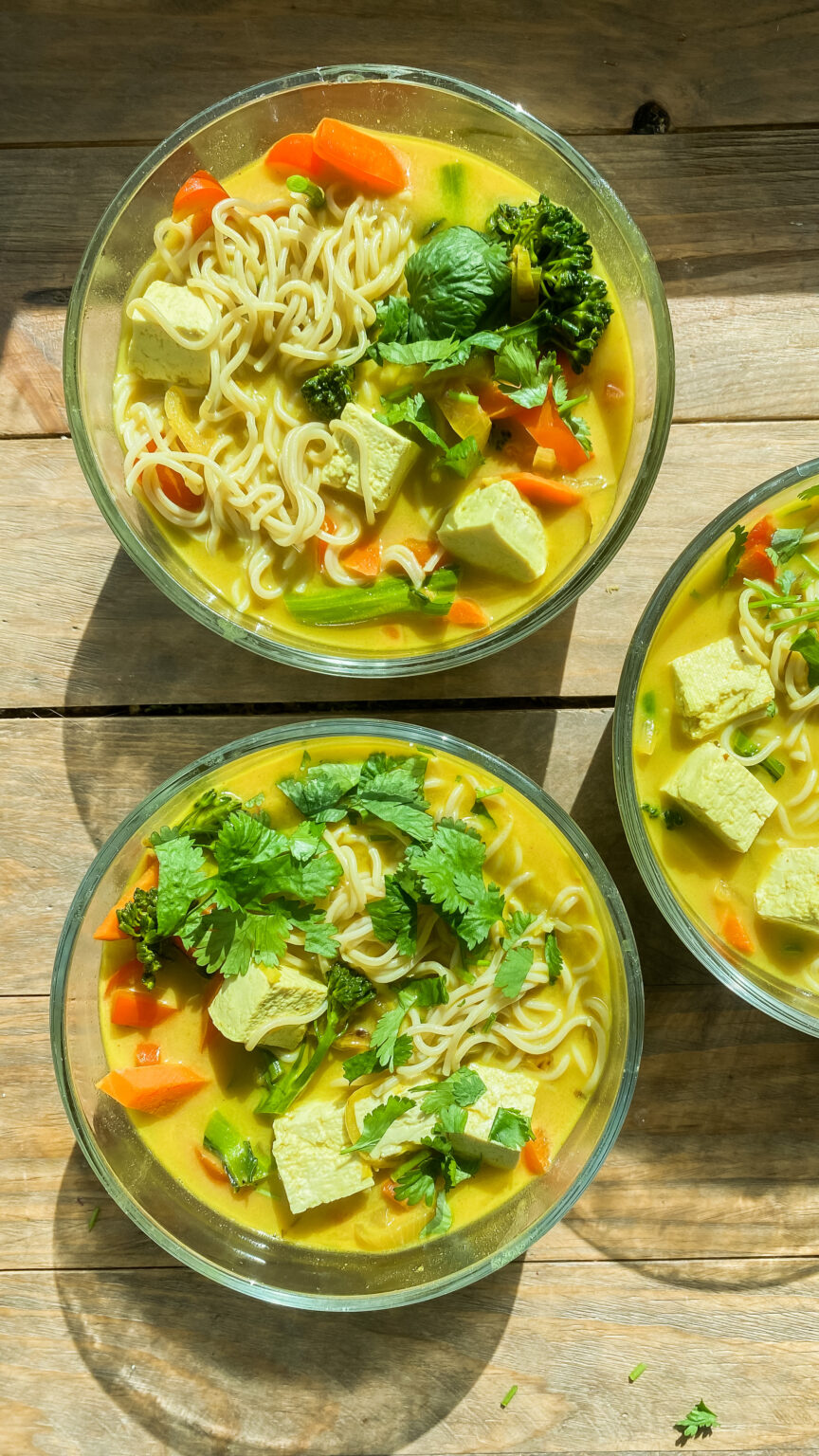 A steaming bowl of tofu curry noodle soup with colorful vegetables like red bell peppers and broccoli, garnished with fresh cilantro, served with udon noodles in a rich coconut curry broth.